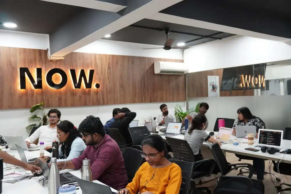 Digital marketing team collaborating in modern office with NOW neon sign, laptops and workspace setup