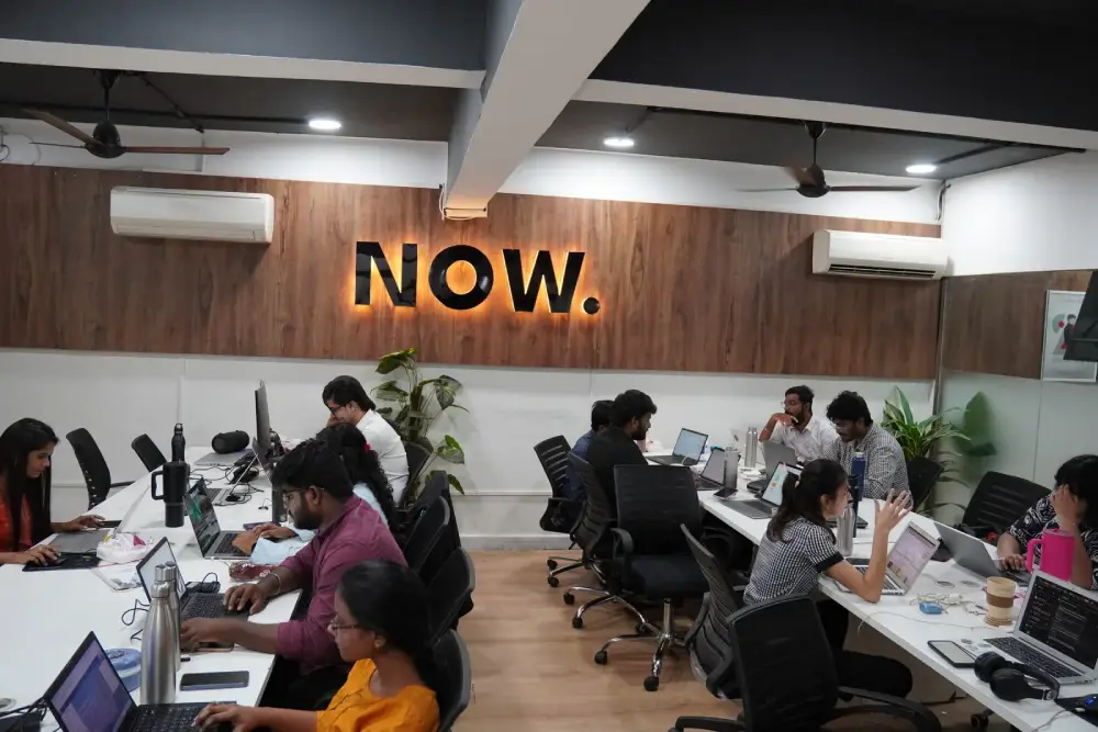 Modern office workspace with diverse team collaborating at desks with computers under NOW neon sign